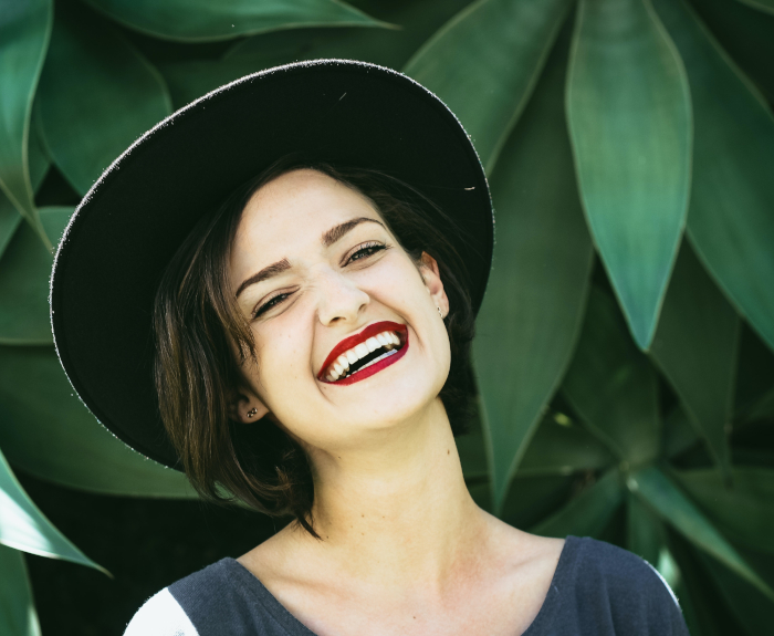 Zahnarztpraxis Dr. Gernot Wagner - Zahnersatz Prothetik - Frau mit Hut und roten Lippen lacht vor grünen Blättern